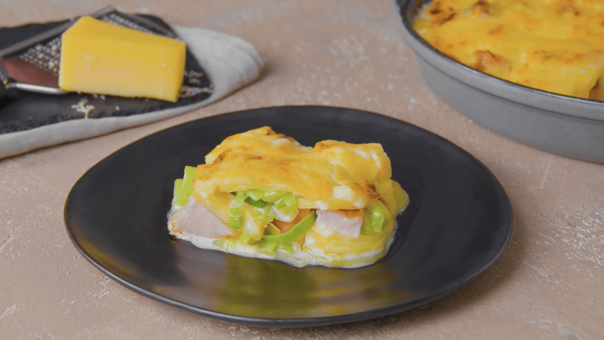 Ein Teller Kasseler-Auflauf mit Lauch und Kartoffeln, dahinter ein Stück Käse mit einer Reibe und die Auflaufform.