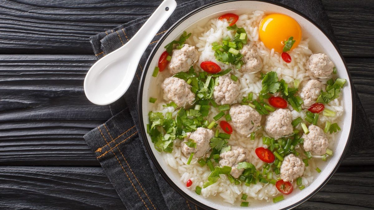 Khao Tom Moo thailändische Reissuppe mit Hackbällchen und Eigelb in weißer Suppenschüssel auf Jeansstoff-Tuch. Daneben weißer asiatischer Suppenlöffel aus Porzelan. Angerichtet auf dunklem Holz. Aufnahme aus der Vogelperspektive.