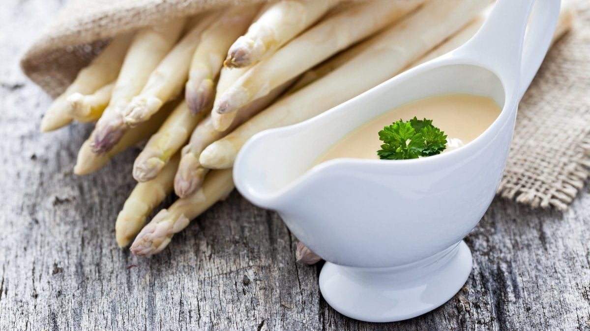 Auf einem Tisch liegen mehrere Stangen weißer Spargel. Daneben steht ein Gefäß aus weißem Porzellan Darin ist eine Sauce hollandaise, eine wichtige Grundsoße.