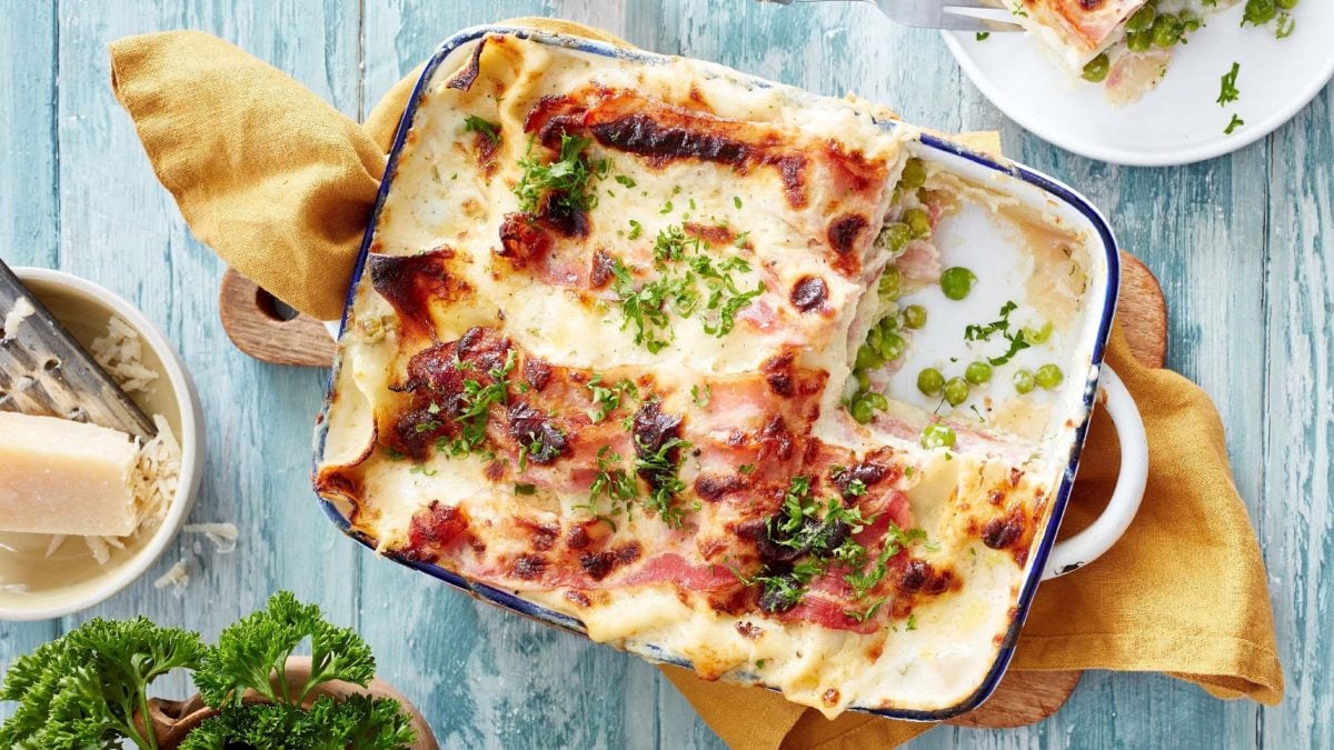 Lasagne alla Carbonara in einer Form mit der cremigen Carbonara-Soße aus Speck, Schmand, Parmesan und Erbsen, serviert auf einem Tuch auf hellem Hintergrund