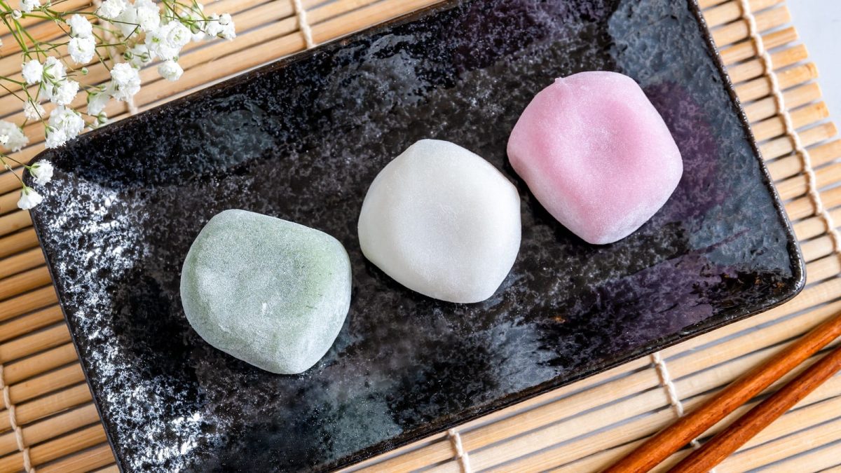 Drei Mochi in verschiedenen Farben, grün, weiß und rosa, auf einem schwarzen, langen Teller, der auf einer Bambusmatte steht, neben einem Strauch weißer Blumen und einem paar Essstäbchen.