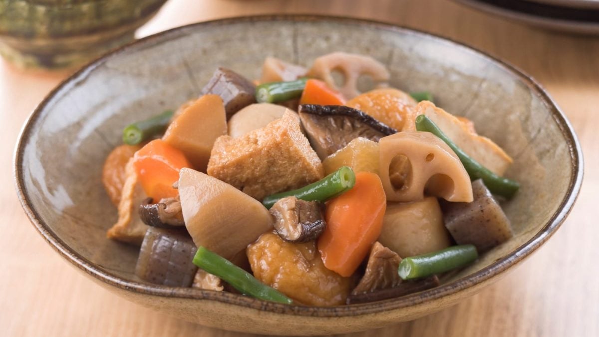 Eine Keramik-Schüssel mit einer Portion des japanischen Gemüse-Eintopfs Nimono aus verschiedenen Wurzeln, Pilzen, Tofu und Bohnen, vor einem Holzhintergrund.