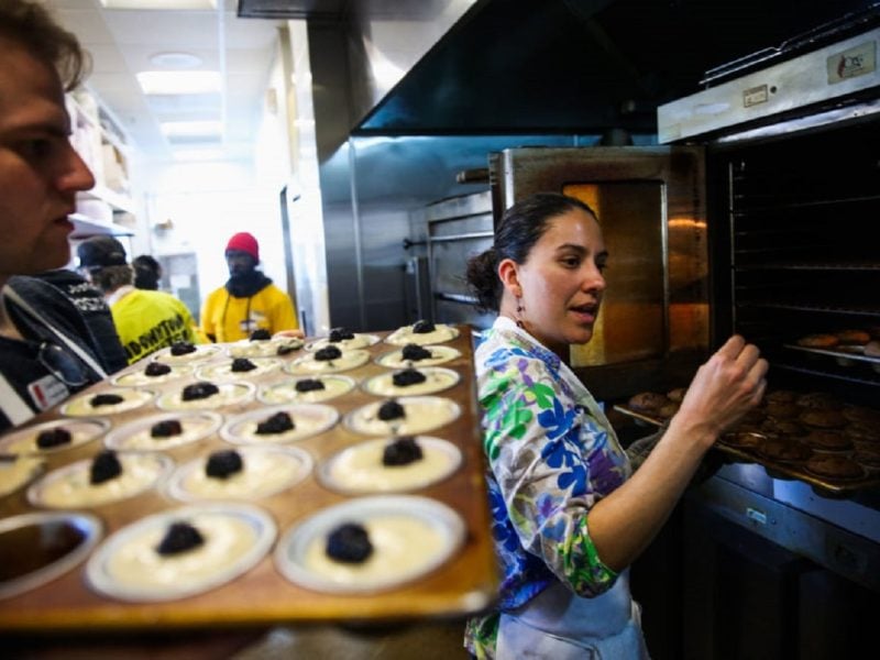 Frau in Profi-Küchen vor geöffnetem Backofen gestikulierend. Mann dahinter hält Backblech Backofen-Zubehör mit Teig. Im Hintergrund Küchenpersonal. Frontalaufnahme.