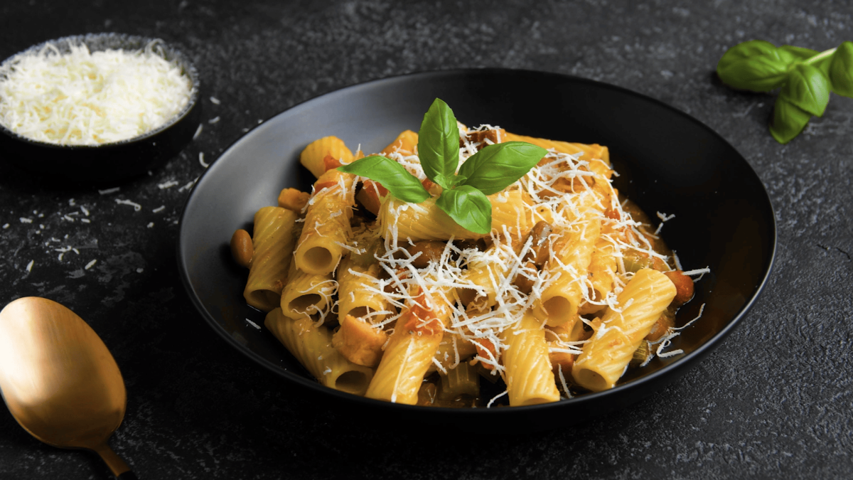 Eine schwarze Schüssel mit einer Portion der Pasta e fagioli mit geriebenem Parmesan bestreut und Basilikumblättern garniert, neben einem Löffel, einer kleinen Schale mit Parmesan und weiteren Basilikumblättern, vor einem dunklen Hintergrund.