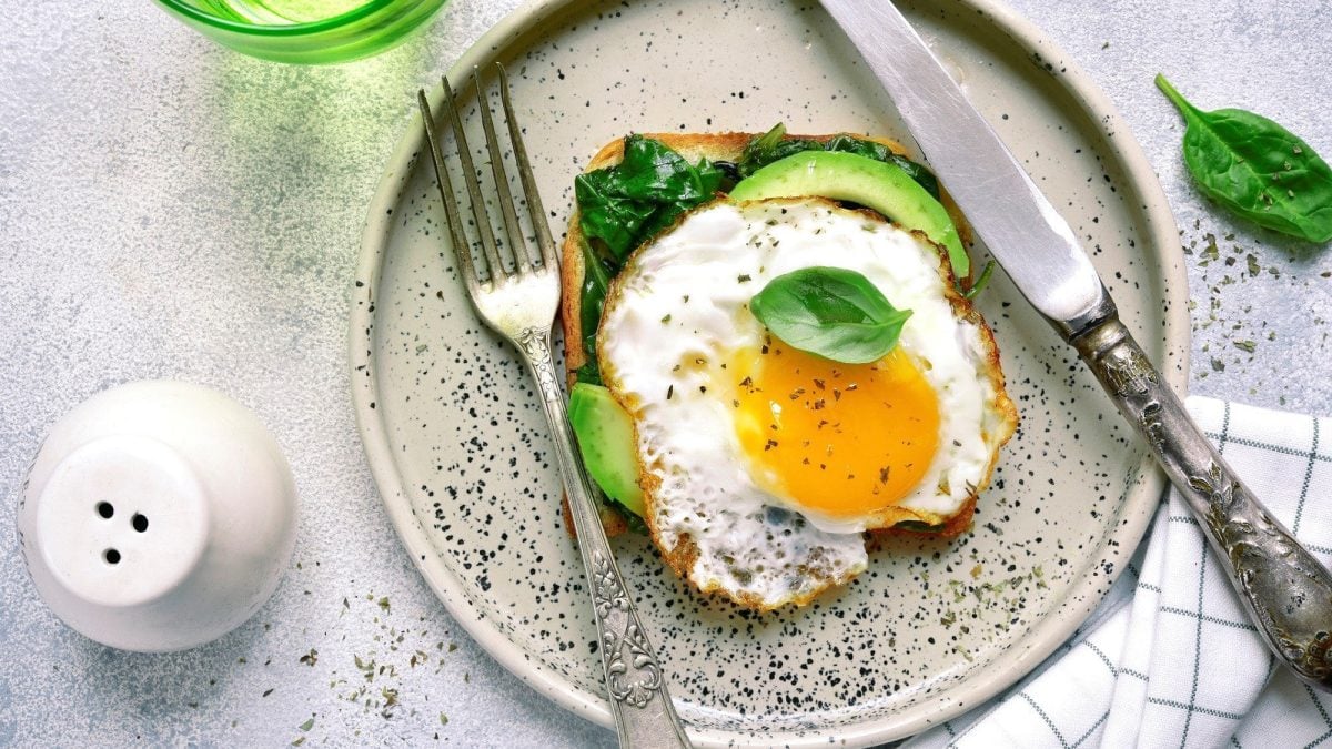 Pesto Eggs auf einem Frühstückstoadt mit Spinat und Avocado. Neben dem Toast liegen Messer und Gabel auf einem gemusterten Teller.