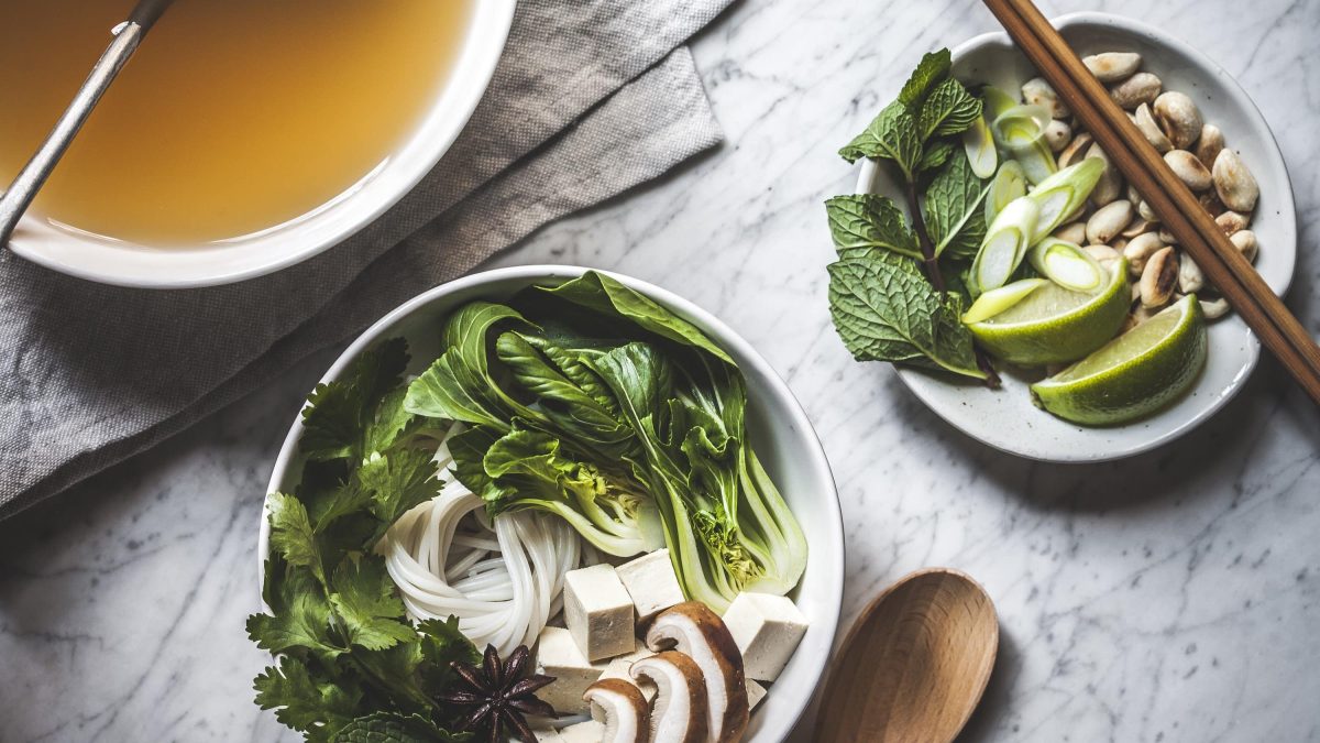 Pho Chay: In einer Schüssel drapierter Pak Choy, Pilze, Bandnudeln, frischer Koreander, daneben ein Teller mit frischer Limette und Erdnüssen und eine Schüssel mit klarer Brühe