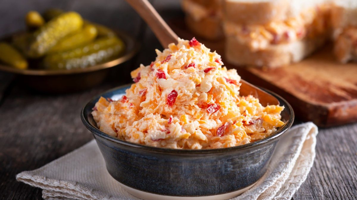 Pimento Cheese Cheddar Chili Dip in schwarzer Schale auf grauem Tuch auf Steintisch. Im Hintergrund Sandwich und saure Gurken in flacher schwarzer Schale. Aufnahme aus der Frontalen.
