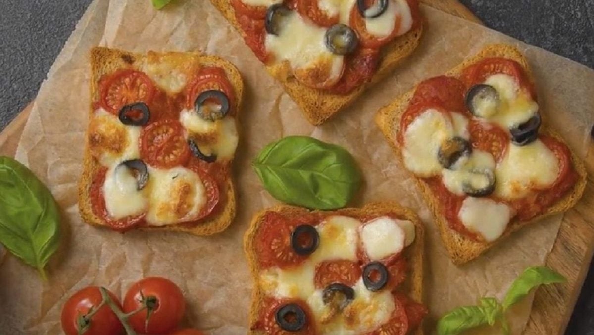 Vier Stücke von unserem Pizza-Toast liegen mit Basilikum und Tomaten garniert auf einem Holzbrett.
