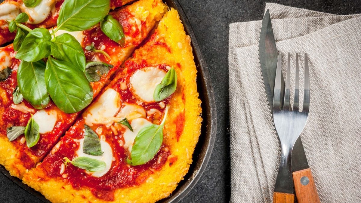 Saftige Polenta-Pizza belegt mit Tomatensoße und Mozzarella, garniert mit frischem Basilikum; serviert in einer heißen Pfanne auf grauem Holzhintergrund