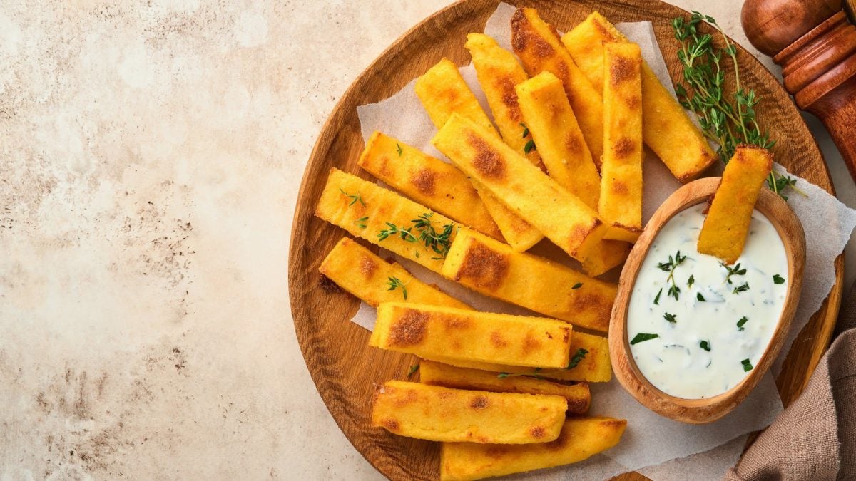 Kross gebackene Polenta-Schnitten mit Kräutern bestreut und dazu frischer Dip serviert auf einem hölzernen Teller auf hellem Hintergrund