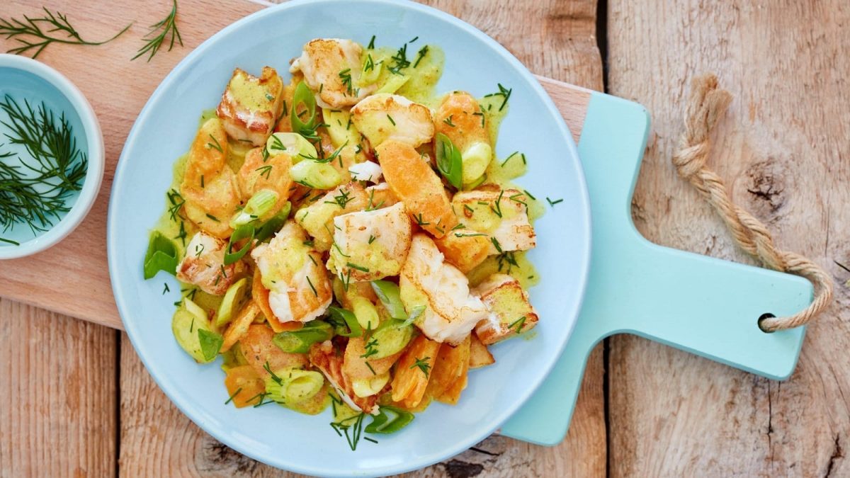 Ein hellblauer Teller auf einem Holz-Schneidebrett, auf dem eine Portion Ragout mit Seelachs und Karotten angerichtet ist, das mit Dill überstreut ist. Neben dem Teller steht eine kleine Schüssel mit Dill. Alles ist vor einem Holzhintergrund von oben fotografiert.
