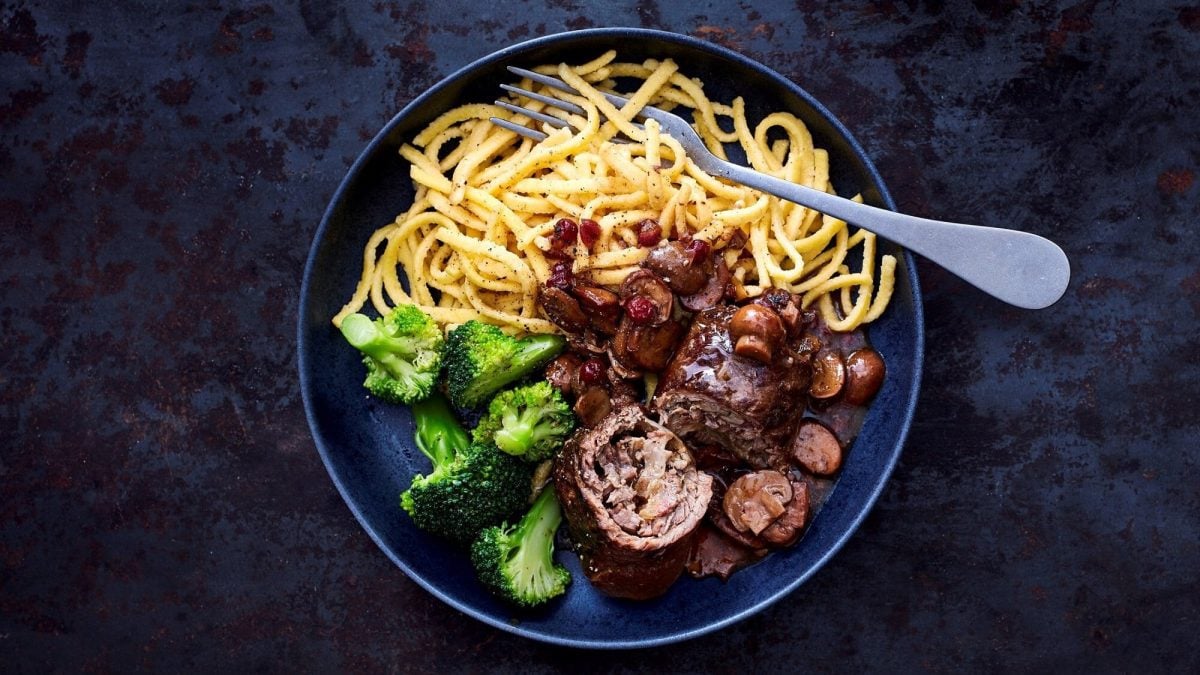 Eine Portion Rinderrouladen in Pilzrahm mit Spätzle auf einem dunkelblauen Teller auf einem dunklen Untergrund. Neben den Spätzle liegt Brokkoli, in den Spätzle liegt eine Gabel.