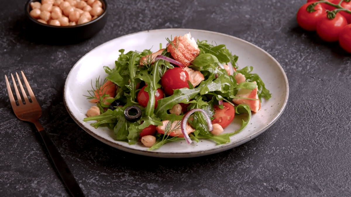 Ein Teller Rucolasalat mit Lachs und Kichererbsen, außerdem Tomaten, roten Zwiebeln und Oliven. Der Teller steht auf dunklem Untergrund, daneben eine Gabel, Tomaten und eine Schale Kichererbsen.