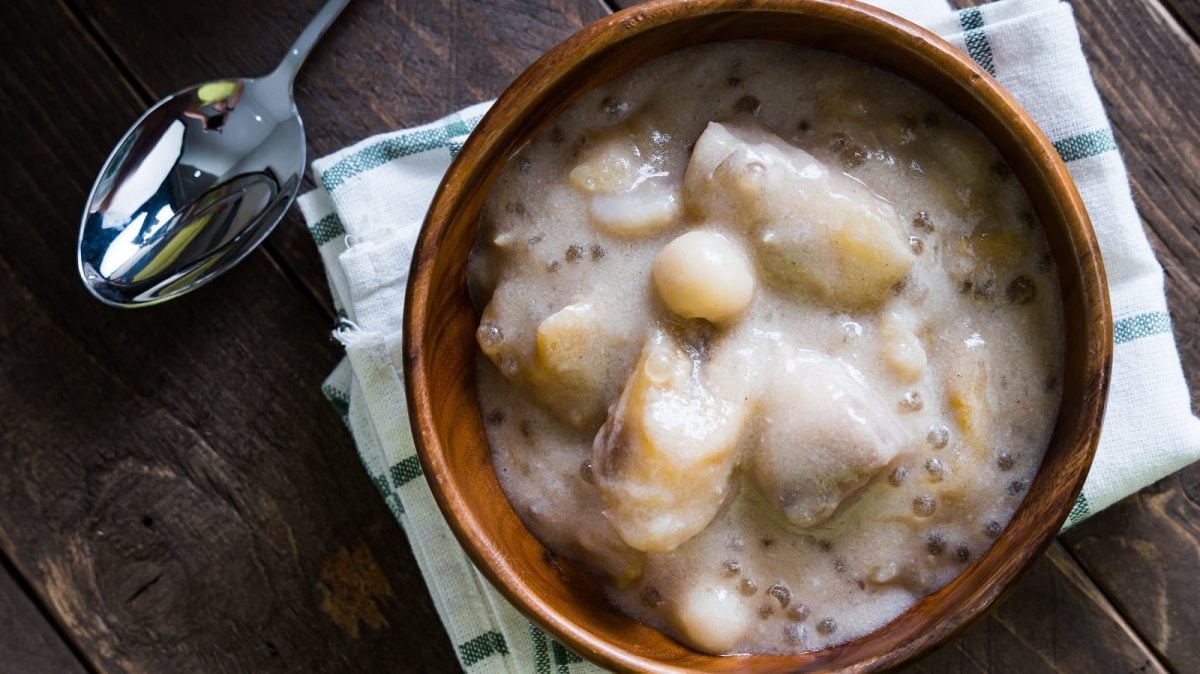 Eine Holzschüssel mit einer Portion der Sago-Suppe mit Taro und Bananen auf einem karierten Tischtuch neben einem Löffel, vor einem dunklen Holzhintergrund von oben.