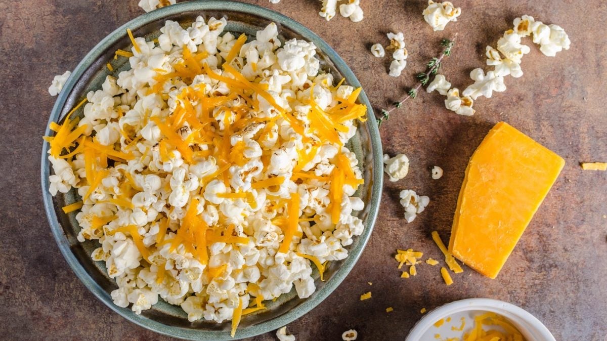 Eine große Schüssel gefüllt mit salzigem Popcorn mit Cheddar Streifen garniert, neben einzelnen Popcorn-Stücken, einem Thymian-Stängel und einem Stück Cheddar, vor einem braunem Hintergrund, von oben.