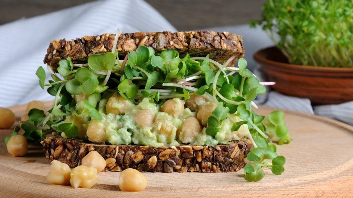 Ein Sandwich mit Kichererbsen-Guacamole und Sprossen auf einem runden Holzbrett. Um das Sandwich herum liegen einige weitere Kichererbsen.