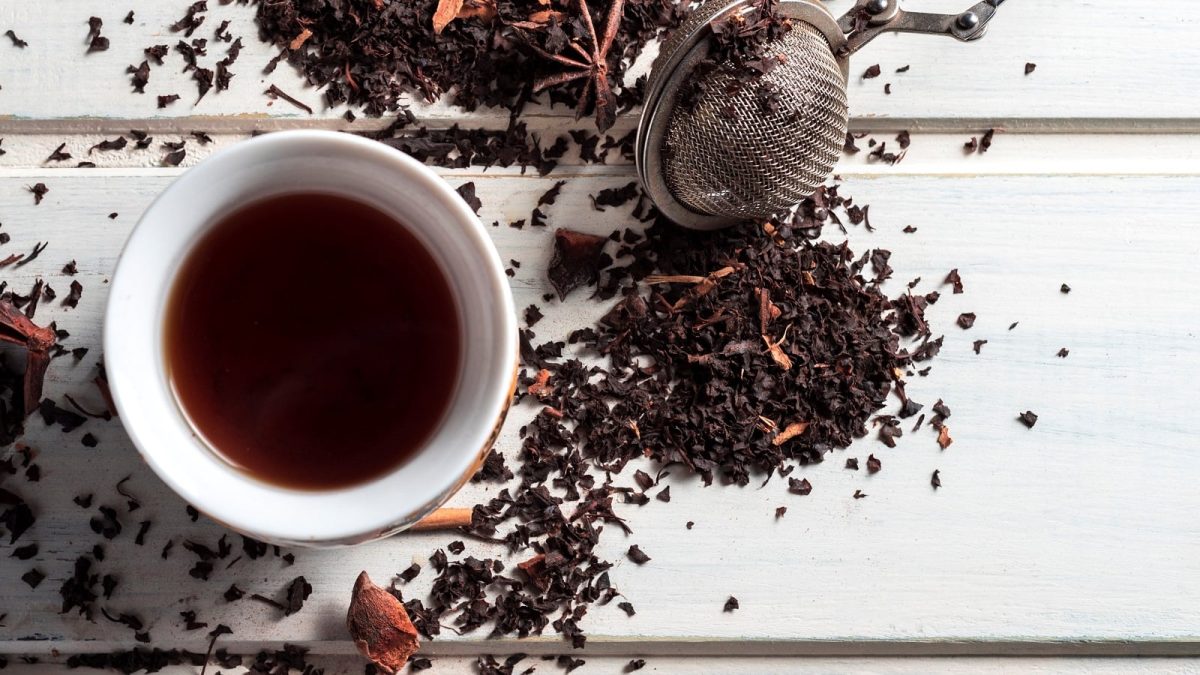 Eine weiße Tasse gefüllt mit dunklem Schwarztee neben einem Teesieb und losem Schwarztee der mit verschiedenen Gewürzen verfeinert ist.