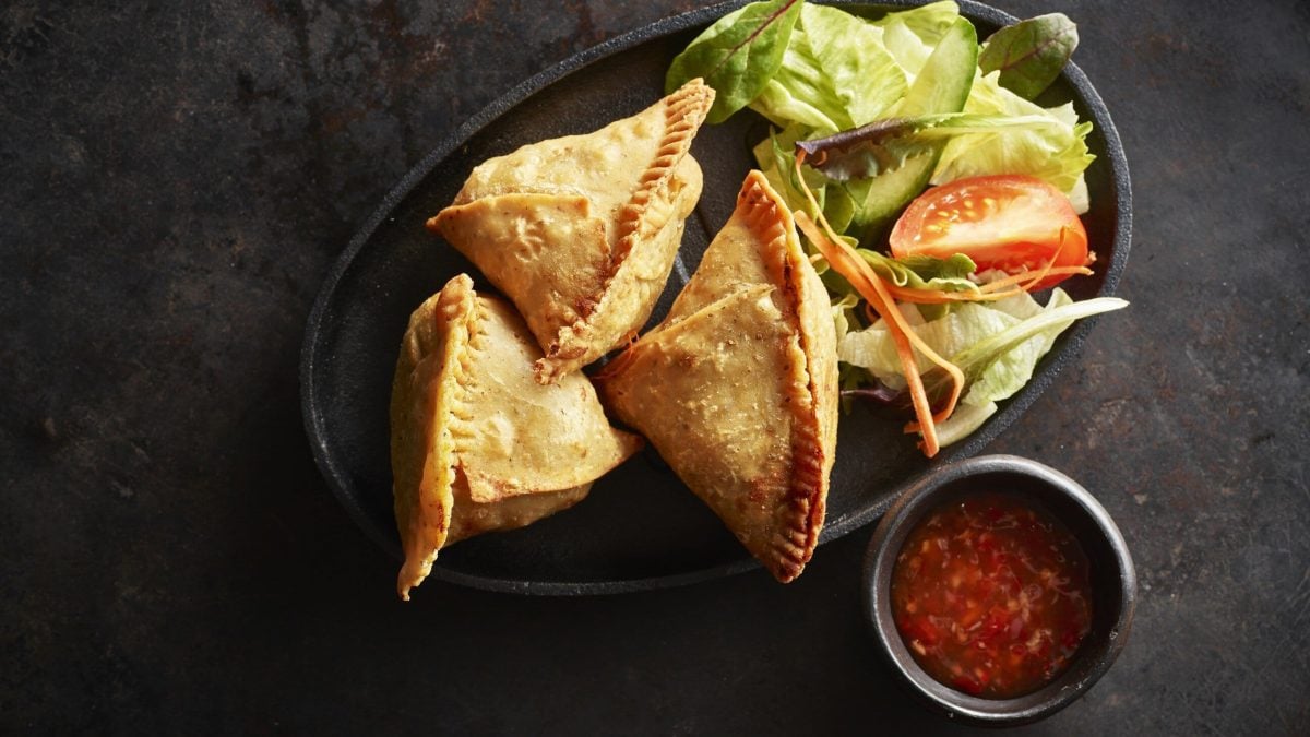 Krosse und würzige selbst gemachte Samosas neben einem Salatbouket, serviert mit Chiliöl auf einer schwarzen Platte auf dunklem Untergrund