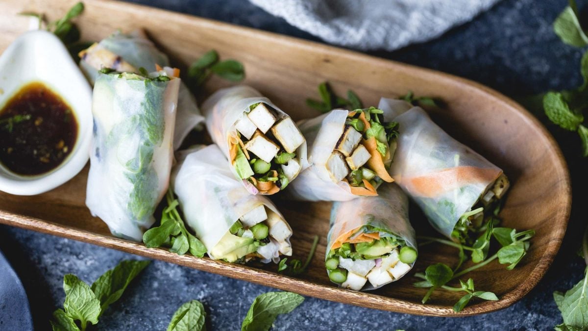 Selbst gemachte Sommer-Rollen, gefüllt mit mariniertem und geröstetem Tofu, Karotte, Spargel, Minze, Koriander und Zwiebeln, serviert mit malziger Sojasauce auf einem hölzernem Brett auf dunklem Untergrund