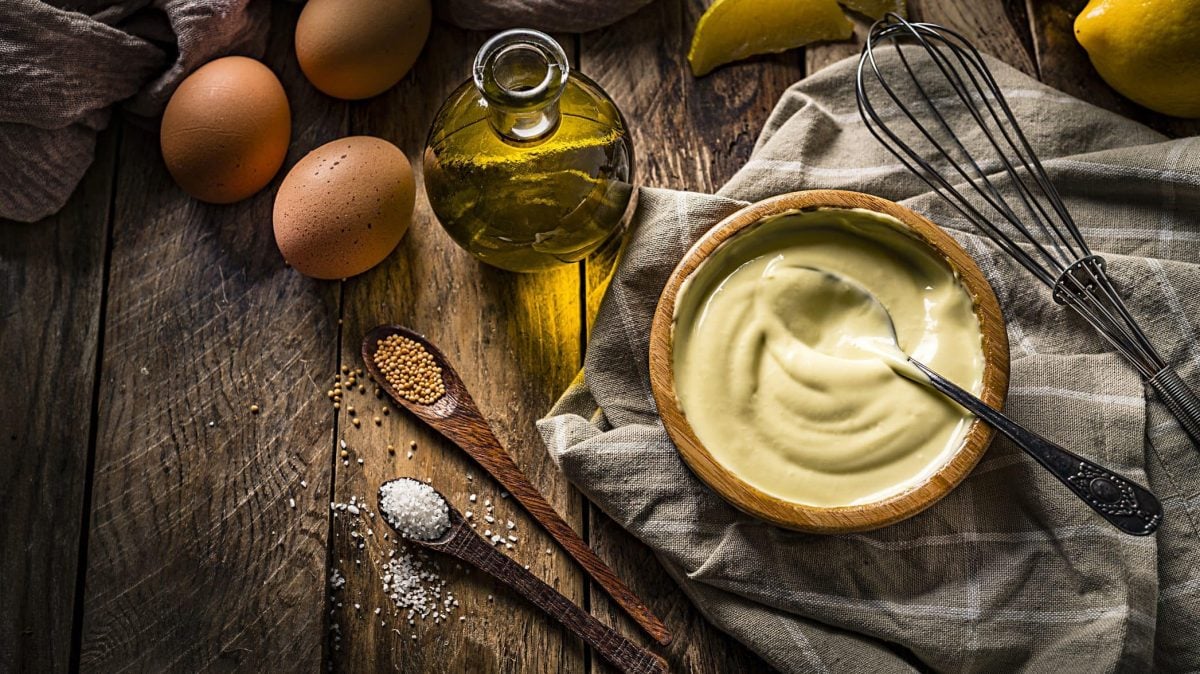 Auf einem Holztisch steht eine Holzschüssel mit selbstgemachter Mayonnaise. Neben ihr liegen ein Schneebesen, Eier, Öl, Salz und Pfeffer.