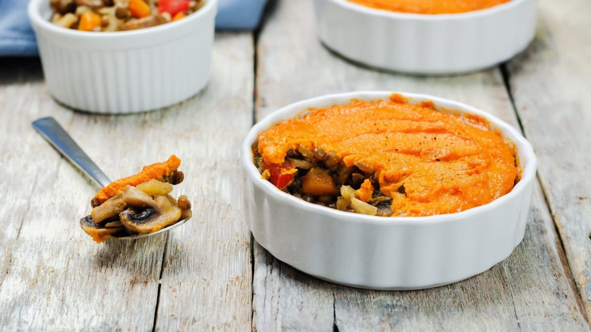 Eine runde Auflaufform Shepherd's Pie mit Süßkartoffelpüree, im Hintergrund sind noch zwei Formen zu erkennen, außerdem liegt ein Löffel mit gekochtem Gemüse daneben.