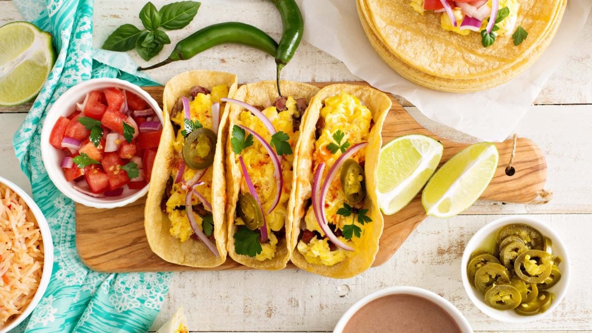 Drei Sin City Breakfast Tacos mit Omelette gefüllt auf Holzbrett . Daneben Tomatensalat. Recht Schälchen mit Jalapenos und Limettenspalten. Peperoni und Koriander als Deko im Bild verteilt. Aufnahme aus der Vogelperspektive.