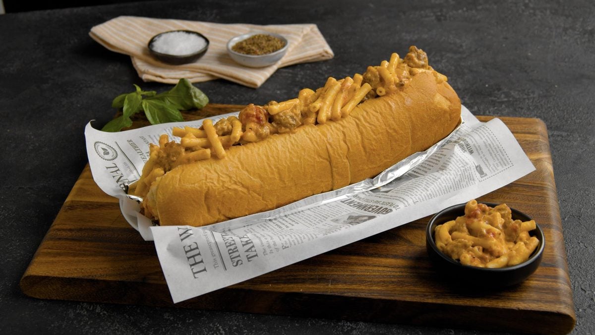 Sloppy Jessica aus "Brookly Nine Nine" Sandwich auf Krepp auf Holzbrett. Daneben Mac and Cheese. Auf dunklem Holztisch oben am Bildrand Creme Fraiche in einer schwarzen Schale. Frontalaufnahme.