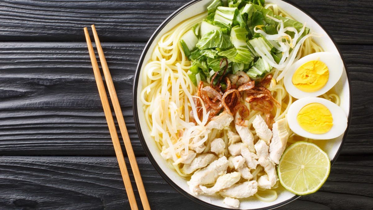 Soto Ayam indonesische Hühnersuppe mit Einlagen in weißer Suppenschüssel mit schwarzem Rand auf schwarzem Holztisch. Daneben Essstäbchen. Aufnahme aus der Vogelperspektive.