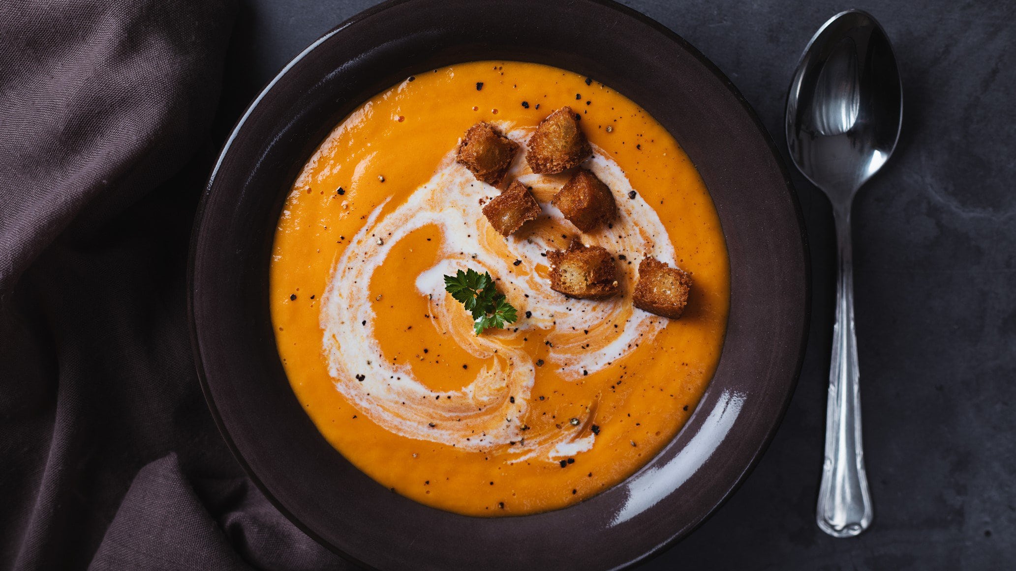 Eine orangefarbene Suppe mit Zimt-Croutons in der Draufsicht, daneben liegt ein Löffel.