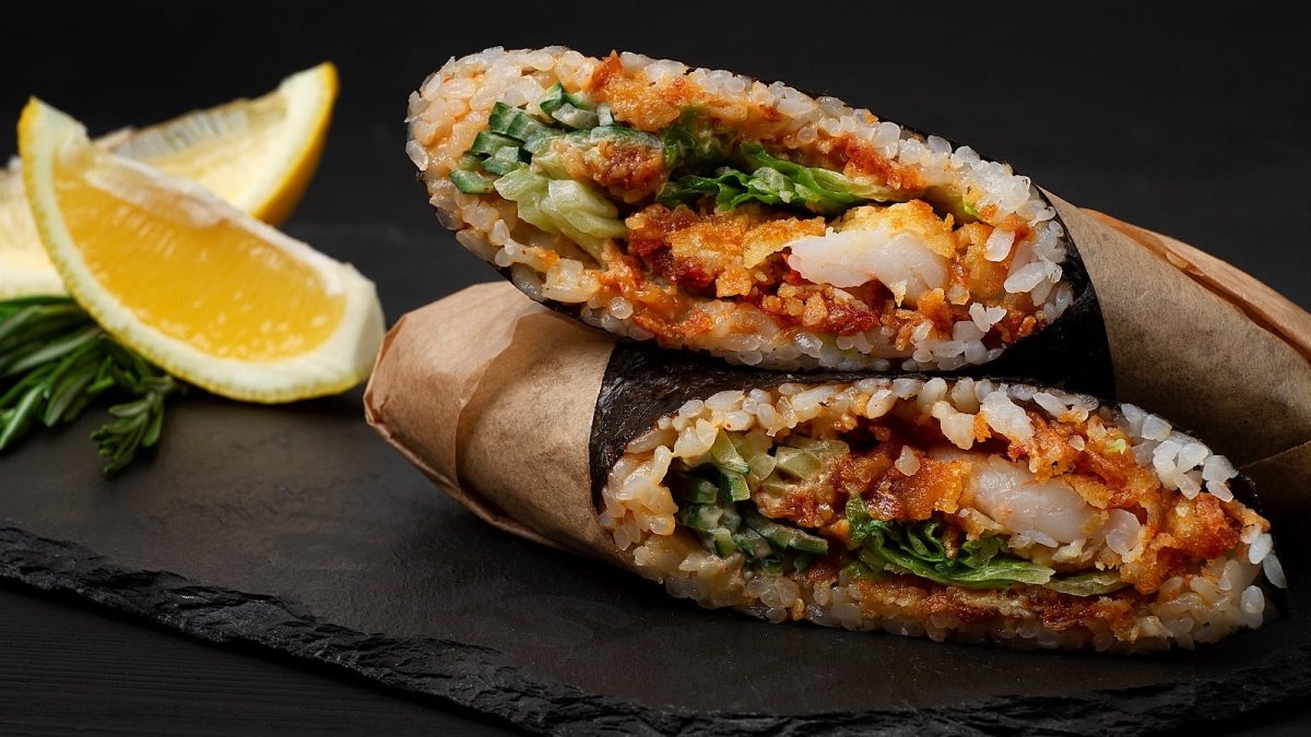 Ein aufgeschnittener Sushi-Burrito mit Shrimps-Tempura und scharfer Sauce in Papier eingewickelt, übereinander gelegen auf einer schwarzen Platte neben Zitronen und Kräutern vor einem schwarzem Hintergrund.