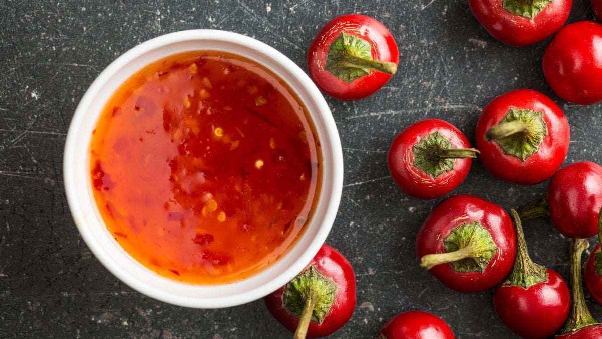 Eine weiße Schale Sweet Chili Sauce auf dunklem Untergrund in der Draufsicht, daneben liegen rote Chilischoten.