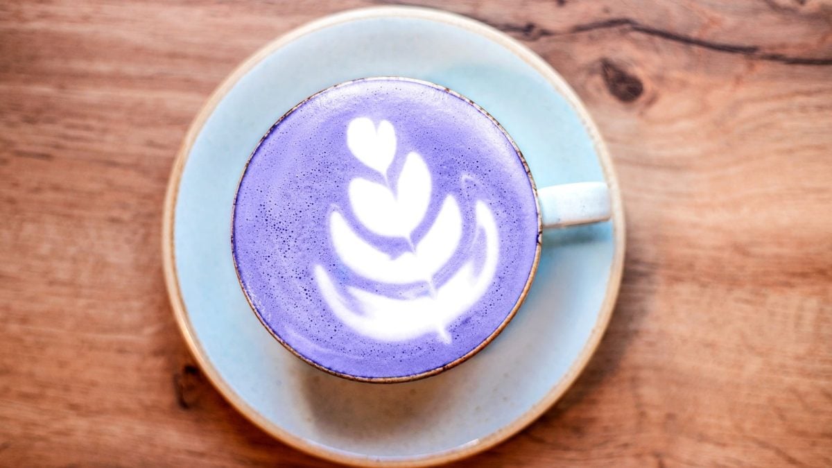 Eine Tasse Taro-Latte mit einer weißen Schaum-Verziehrung, auf einer Untertasse stehend, vor einem Holzhintergrund, von oben.