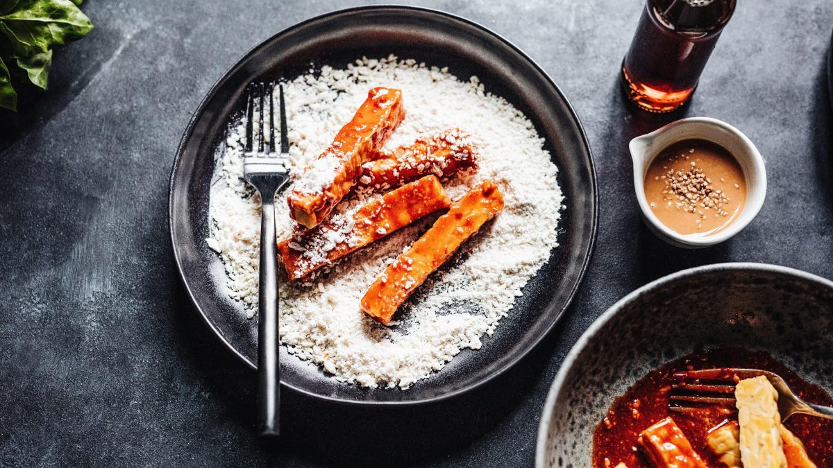 Tempeh-Fritten in der Zubereitung, mariniert und in Mehl gewendet, bereit frittiert zu werden. Daneben stehen Erdnusssauce und Fischsauce bereit, auf dunklem Hintergrund
