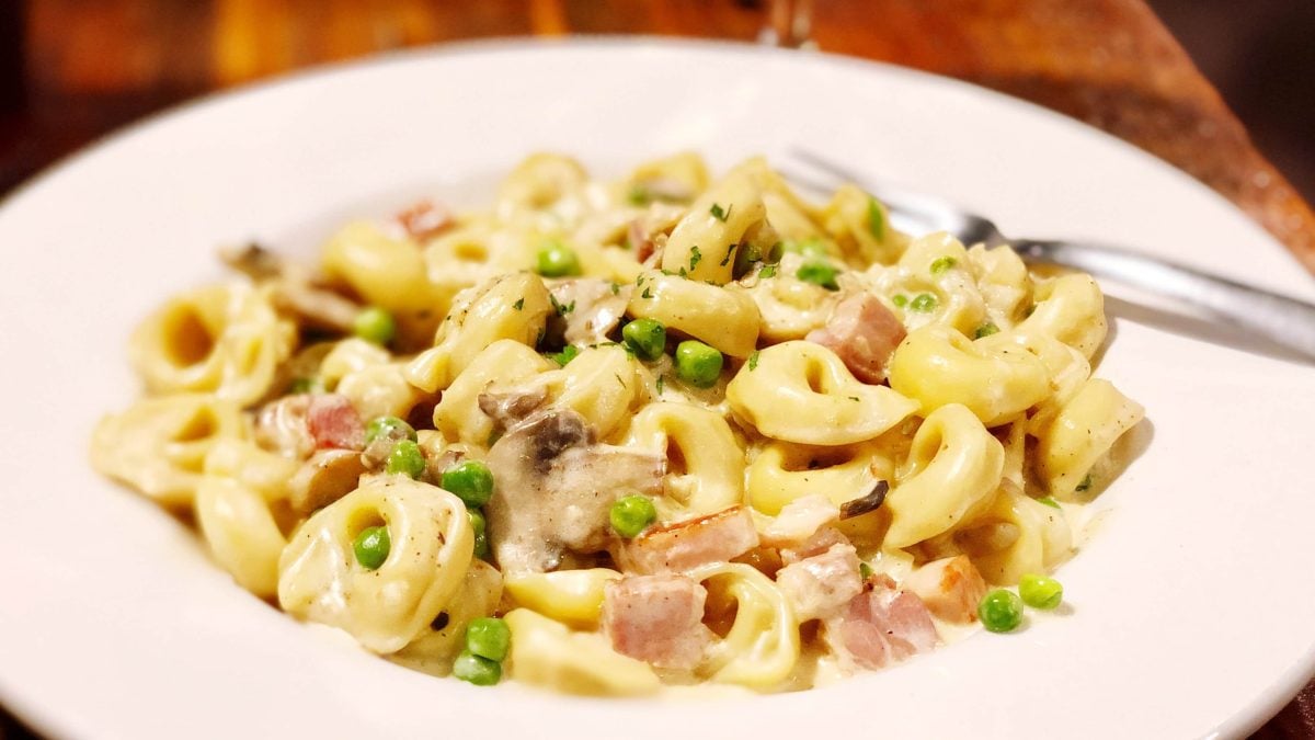 Tortellini à la creme aus Das große Fressen auf weißem Pastateller und dunkelbraunem Tisch. Rechts eine Gabel auf dem Teller. Aufnahme aus der Draufsicht.
