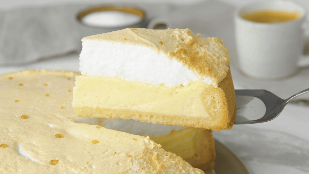 Ein Stück Tränenkuchen wird mit Hilfe eines Kuchenhebers von dem Rest des auf einer Kuchenplatte stehenden Tränenkuchens gehoben, sodass man die Haube, den Belag und den Boden von der Seite sehen kann. Im Hintergrund ist eine Tasse Kaffee sowie Zucker zu sehen vor einem grauen Hintergrund.