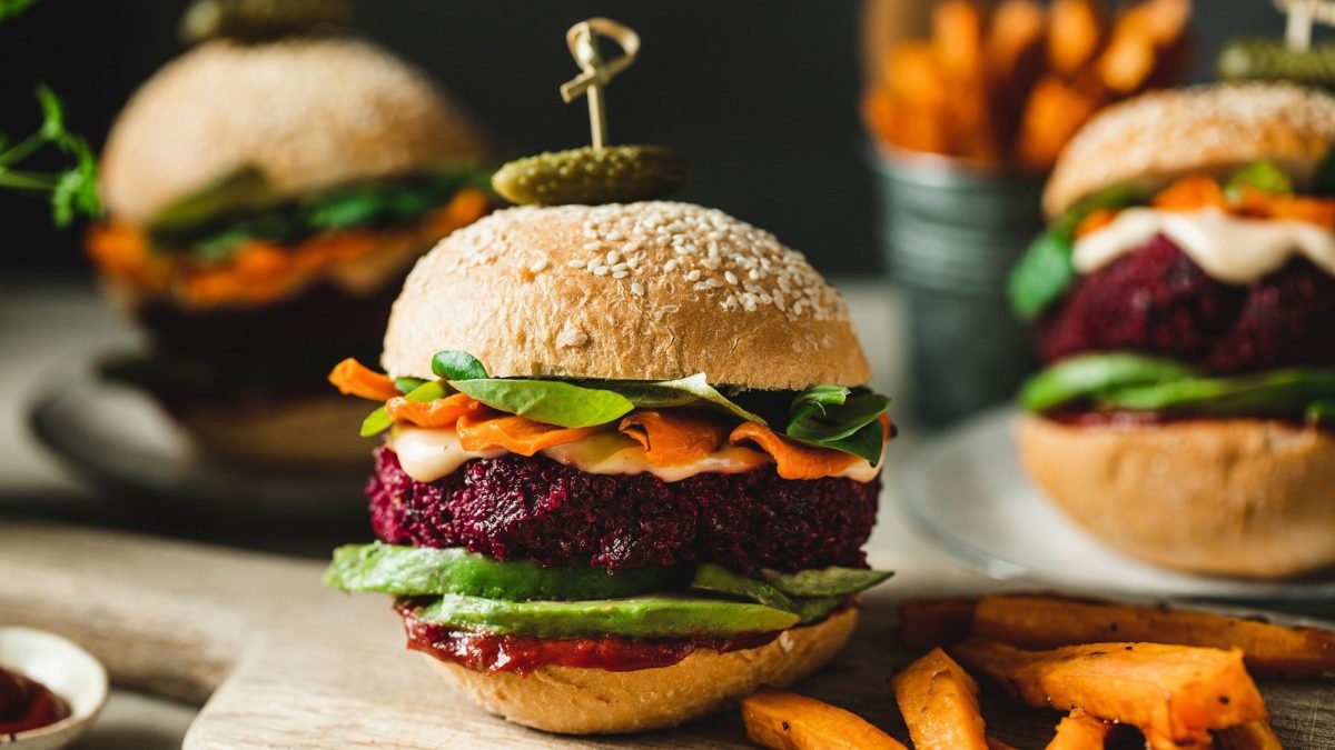 Vegane Burger mit Rote-Bete-Patty und Avocado liegen auf einem hellen Holzbrett. Die Burger sind zusätzlich angerichtet mit Mayonnaise, BBQ-Sauce, Karotten und Salat
