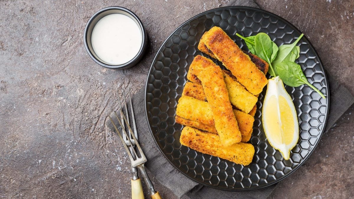 Vegane Fischstäbchen auf einem schwarzen Teller mit zitrone und Spinatblättern serviert. Daneben ein Schälchen mit Mayonnaise auf einem steinernen Untergrund.