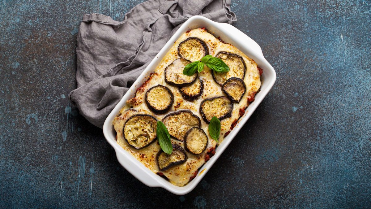 Eine Auflaufform vegane Moussaka auf dunklem Untergrund in der Draufsicht, daneben ein graues Geschirrtuch.