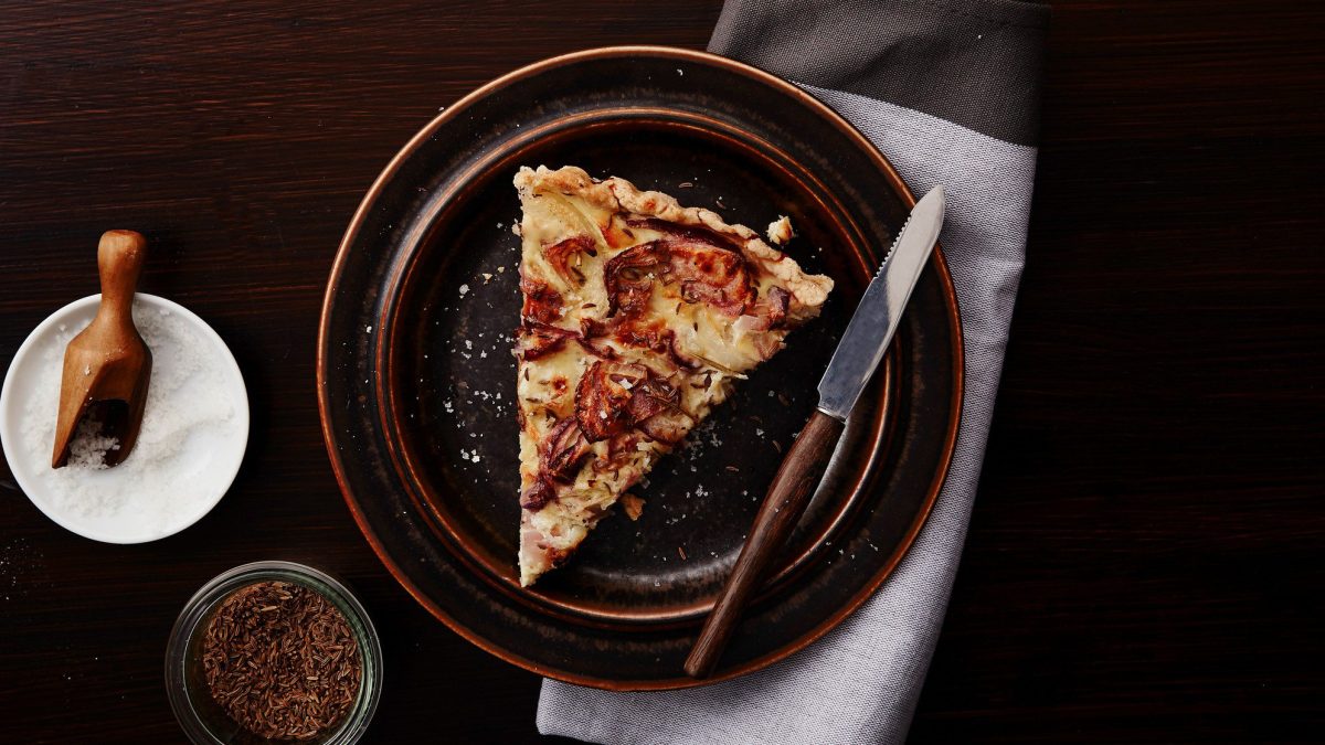 Ein Teller vegane Quiche Lorraine mit einem Messer auf dunklem Untergrund in der Draufsicht. Daneben eine Schale mit Salz und Kümmel.