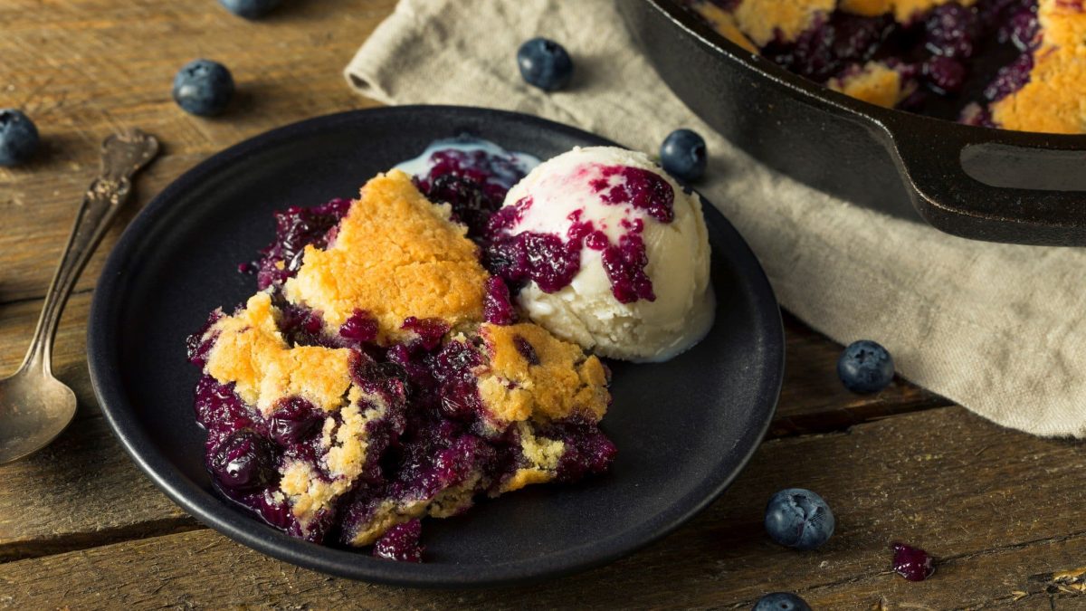 Ein Teller veganer Heidelbeerauflauf mit einer Kugel Vanilleeis, daneben liegen ein Löffel und einige Heidelbeeren verstreut.