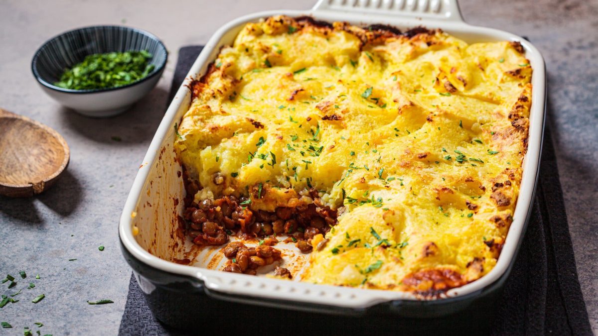 Eine Auflaufform mit veganem Shepherd's Pie, aus der schon ein Stück fehlt, daneben eine Schale mit Kräutern.