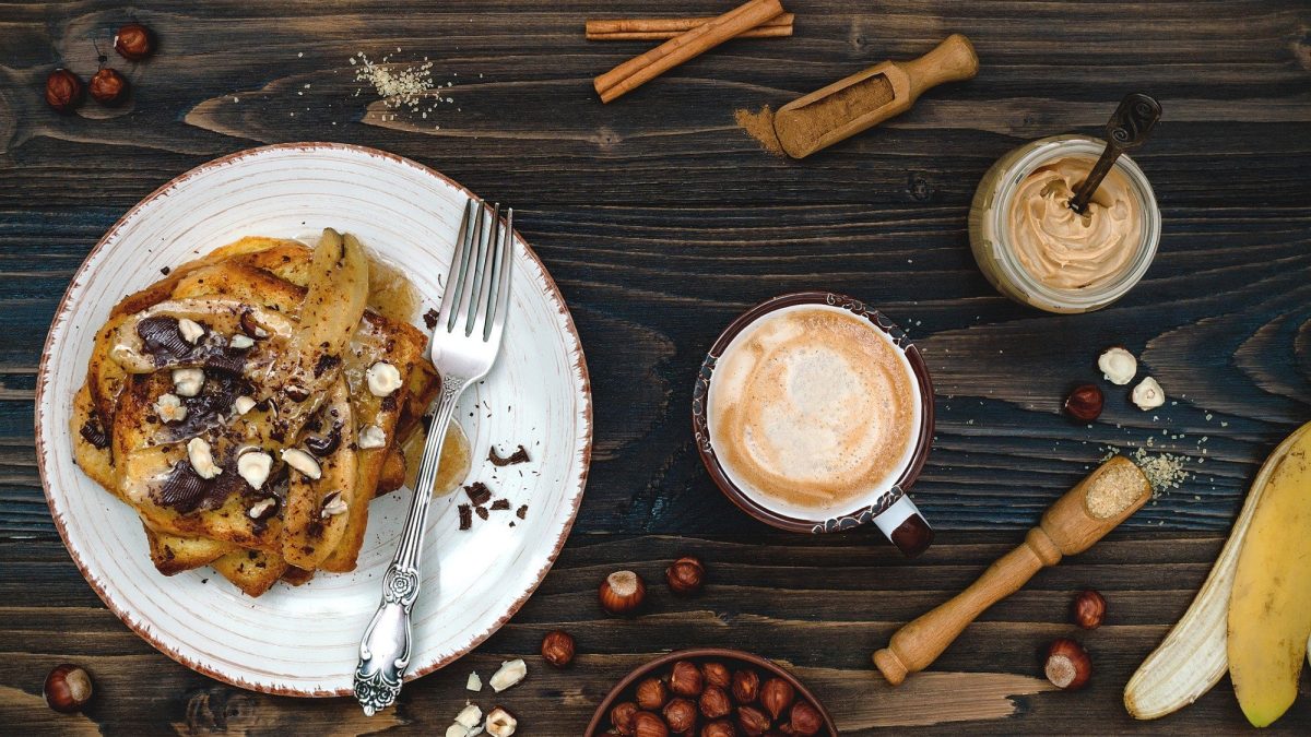 Unser veganes French Toast mit gebackenen Bananen liegt auf einem weißen Teller. Das French Toast ist angerichtet mit Schokolade und Haselnüssen.