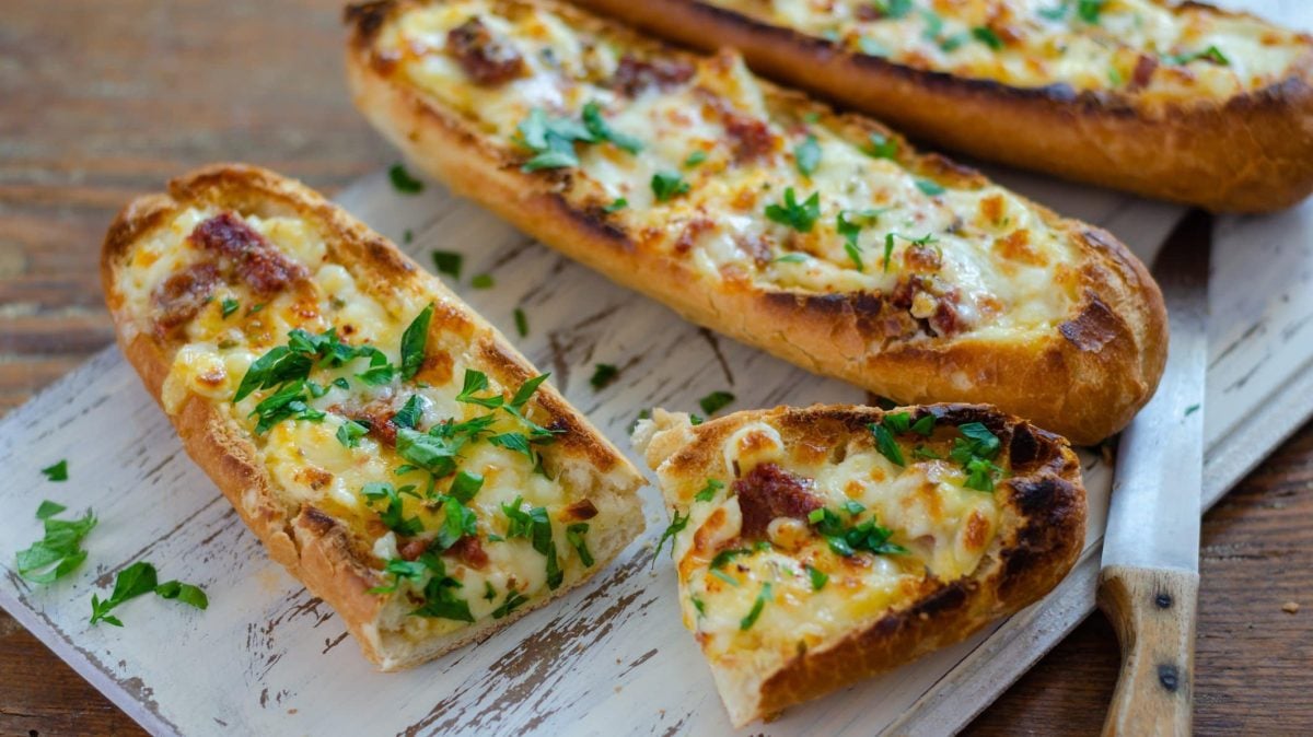 Vegane Ofenbaguettes liegen auf einem Holzbrett, daneben ein Messer.