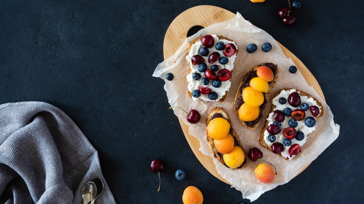 Drei Scheiben mit vitaminreichem Frühstücksbrot liegen auf einem ovalen Tablett auf einem dunklen Untergrund. Die Brote sind angerichtet mit Hüttenkäse, Schokocreme, Aprikosen, Kirschen und Heidelbeeren. Ein wirklich gesundes Frühstück.