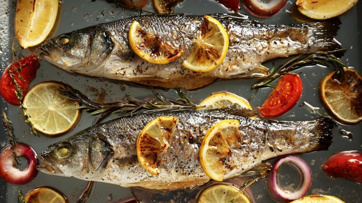 Aus der Vogelperspektive: Zwei Fische, es handelt sich um Wolfsbarsche, liegen auf einem Blech. Drum herum liegen Tomaten und Zitronenschieben sowie Rosmarin und Thymian.