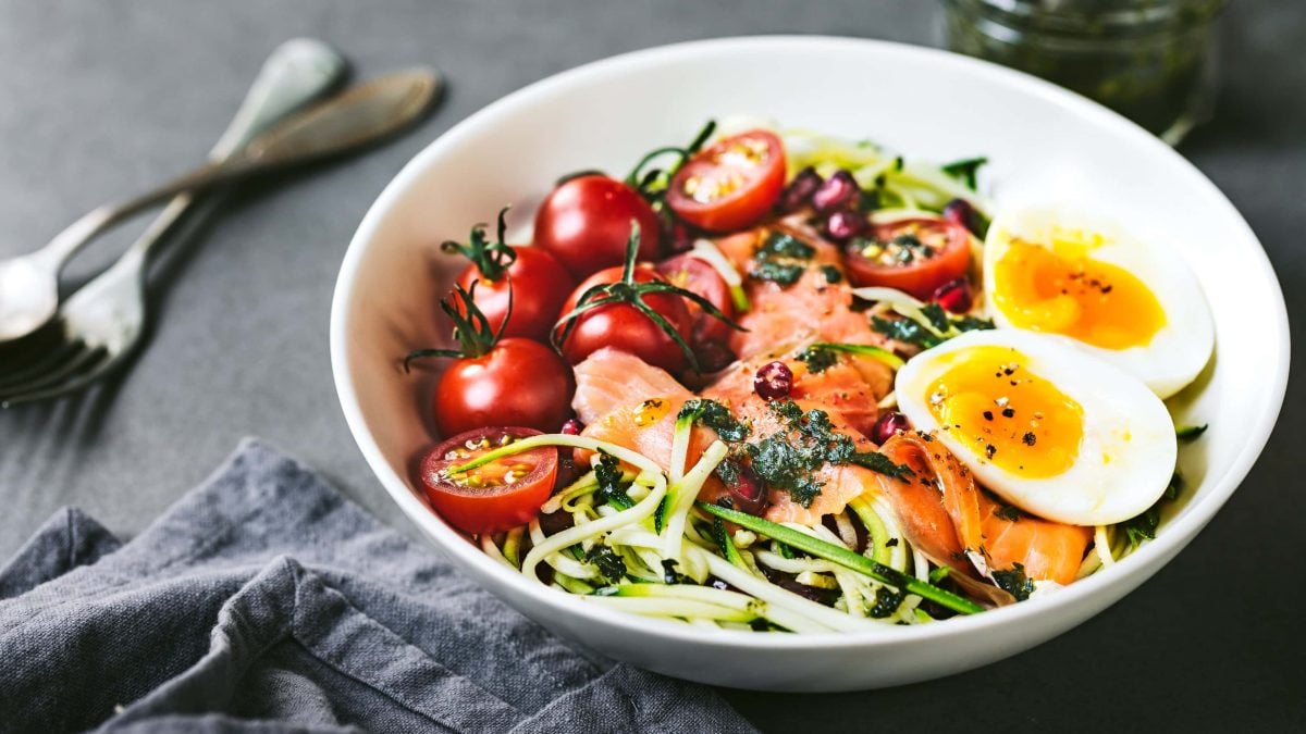 Zoodle-Bowl Low Carb mit Ei, Tomate, Lachs in weißer Schale auf grauem Tisch. Daneben graue Serviette. Oben link zwei Gabeln. Draufsicht.