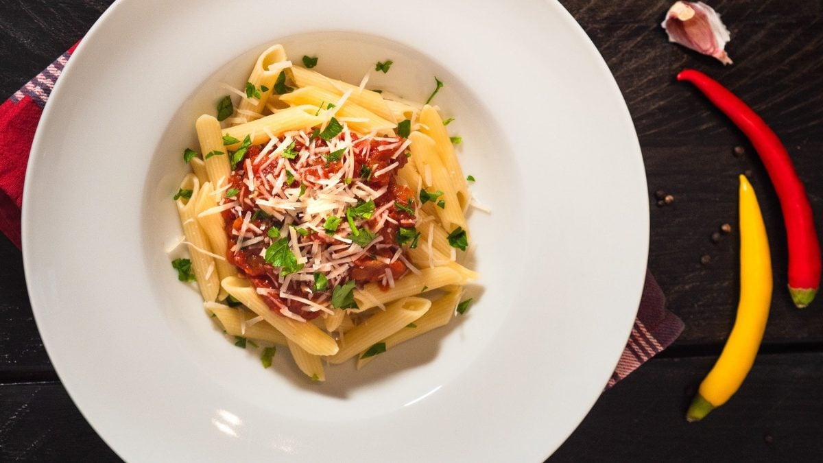 Penne all'arrabiata auf weißem Teller bestreut mit Pecorino und Petersilie auf dunklem Untergrund.