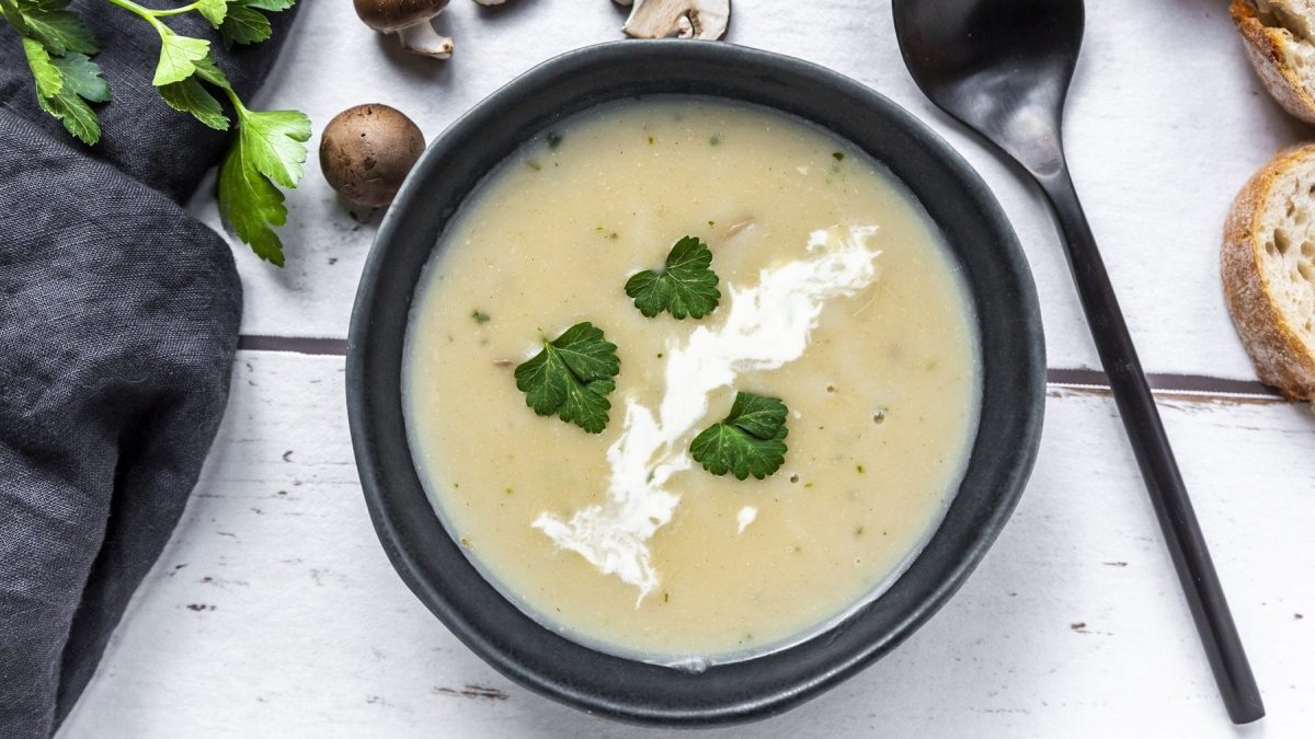 Aus der Vogelperspektive: Auf einem Tisch steht eine cremige Champignonsuppe in einer schwarzen Schale. Daneben liegen Persilienzweige, ein Löffel und einige Scheiben Weißbrot.