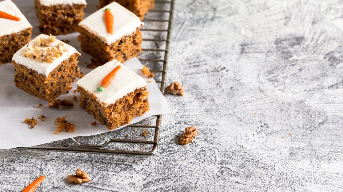 Auf einem Backgitter liegen einige Stück Karottenkuchen in Quadrate geschnitten. Sie sind dekoriert mit Frischkäse, Walnüssen und Marzipan-Karotten.