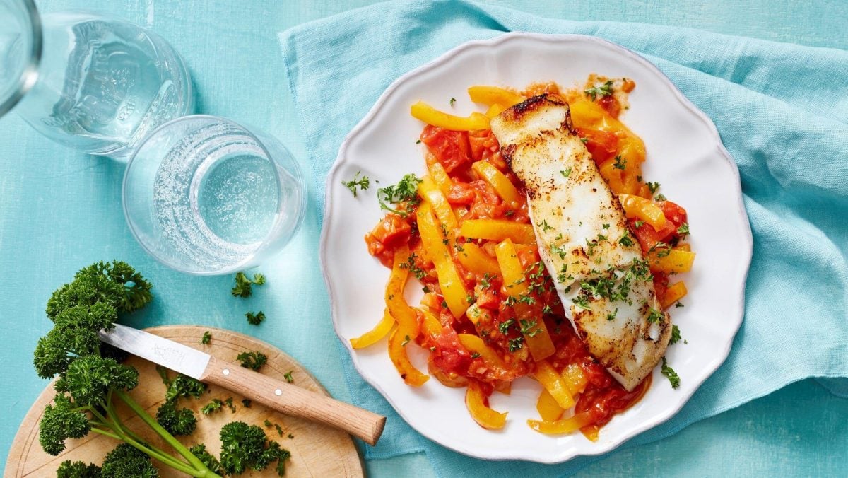 Fischfilet auf Paprikagemüse in Draufsicht mit Glas Petersilie und blauer Tischdecke